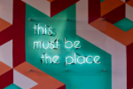 Neonschild mit der Aufschrift „this must be the place“ auf einer bunten geometrischen Wand.